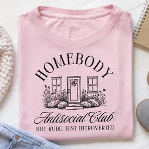 May include: Pale pink t-shirt featuring the text "HOMEBODY Antisocial Club" in black, with a house graphic. The design includes the phrase "NOT RUDE, JUST INTROVERTED". A casual, comfortable top.