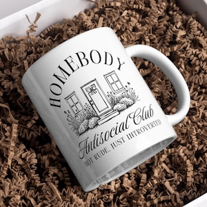 May include: White ceramic mug with the text "HOMEBODY Antisocial Club HOT RUDE, JUST INTROVERTED" and a house illustration. The mug is inside a white box filled with brown paper shreds.