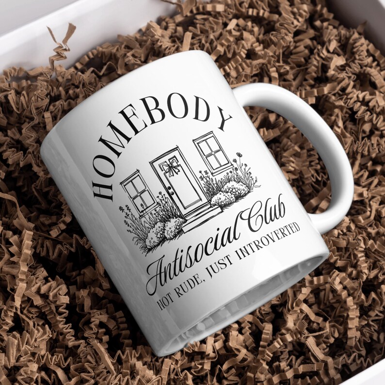 May include: White ceramic mug with the text "HOMEBODY Antisocial Club HOT RUDE, JUST INTROVERTED" and a house illustration. The mug is inside a white box filled with brown paper shreds.