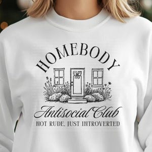 May include: White sweatshirt featuring a black graphic. The design includes the word "HOMEBODY" above a house illustration, with "Antisocial Club" below. The text "NOT RUDE, JUST INTROVERTED" is printed beneath.