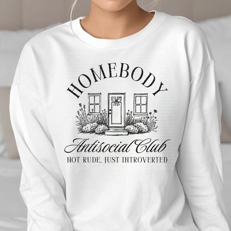 May include: White long-sleeved sweatshirt with a black graphic. The graphic shows a house illustration with the words "HOMEBODY" above and "Antisocial Club" below. The text "NOT RUDE, JUST INTROVERTED" is also included.