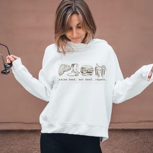 May include: White sweatshirt with a graphic of a steak, cowboy boot, burger, and saddle. The text "raise beef. eat beef. repeat." is printed below the graphics. The sweatshirt has long sleeves and a hood.