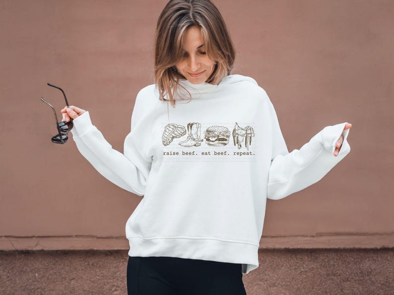 May include: White sweatshirt with a graphic of a steak, cowboy boot, burger, and saddle. The text "raise beef. eat beef. repeat." is printed below the graphics. The sweatshirt has long sleeves and a hood.
