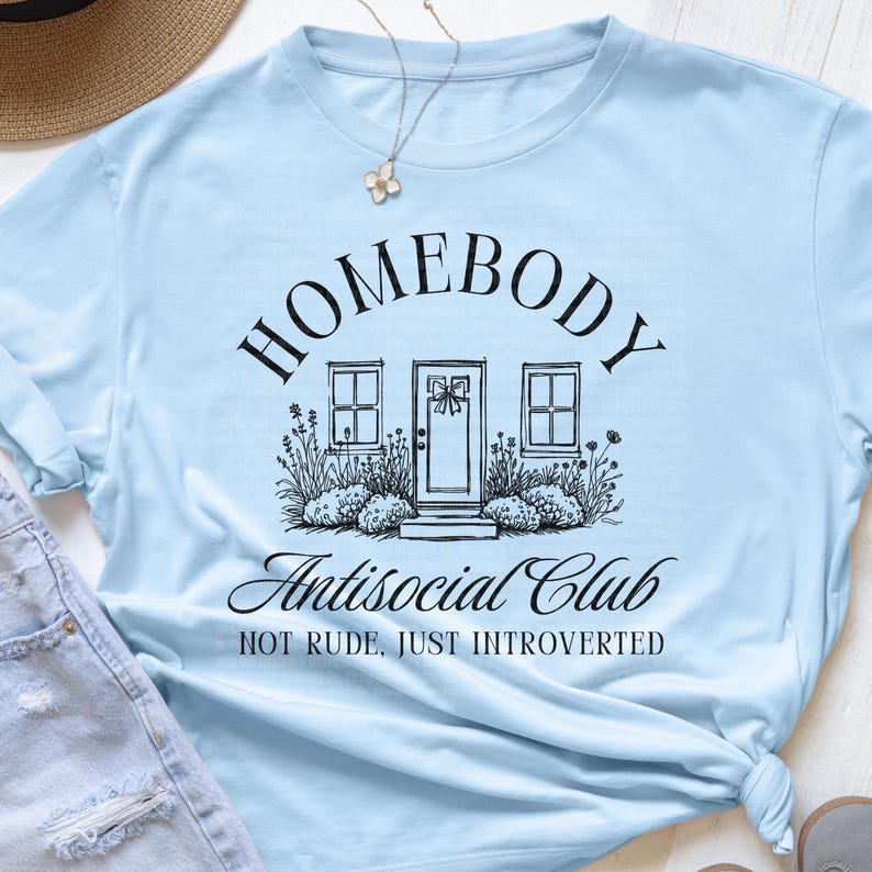 May include: Pale blue t-shirt featuring the text "HOMEBODY Antisocial Club" in black script, with a house illustration. The design includes the text "NOT RUDE, JUST INTROVERTED" below the main text. A casual, everyday garment.