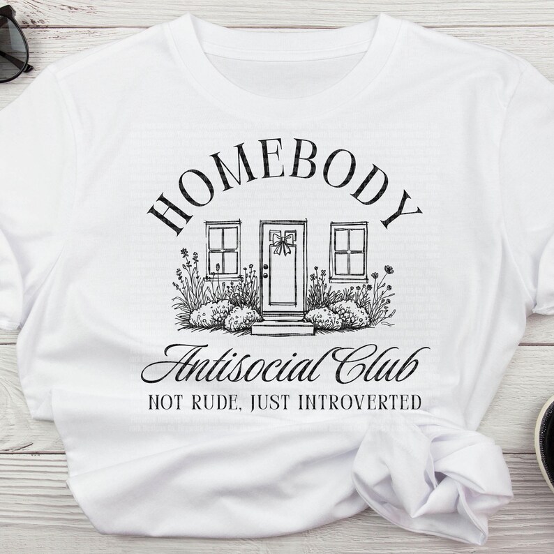May include: White t-shirt featuring a black illustration of a house with the words "HOMEBODY" and "Antisocial Club". The text "NOT RUDE, JUST INTROVERTED" is also included. The shirt is made of a soft material and is suitable for casual wear.
