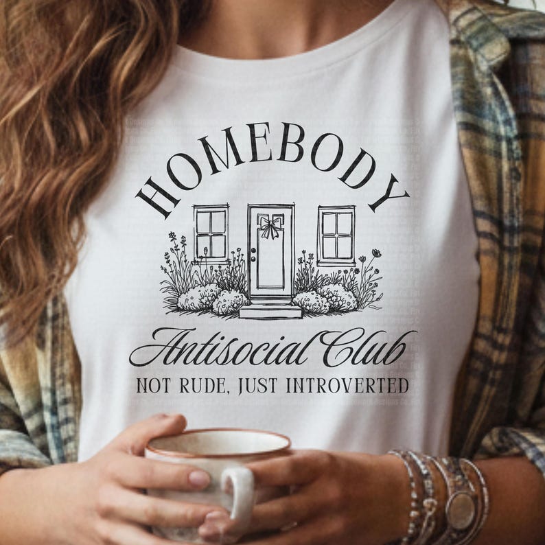 May include: White t-shirt featuring a black illustration of a house with the words "HOMEBODY Antisocial Club" above and below the house graphic. The text "NOT RUDE, JUST INTROVERTED" is below the club name. A person is holding a mug.