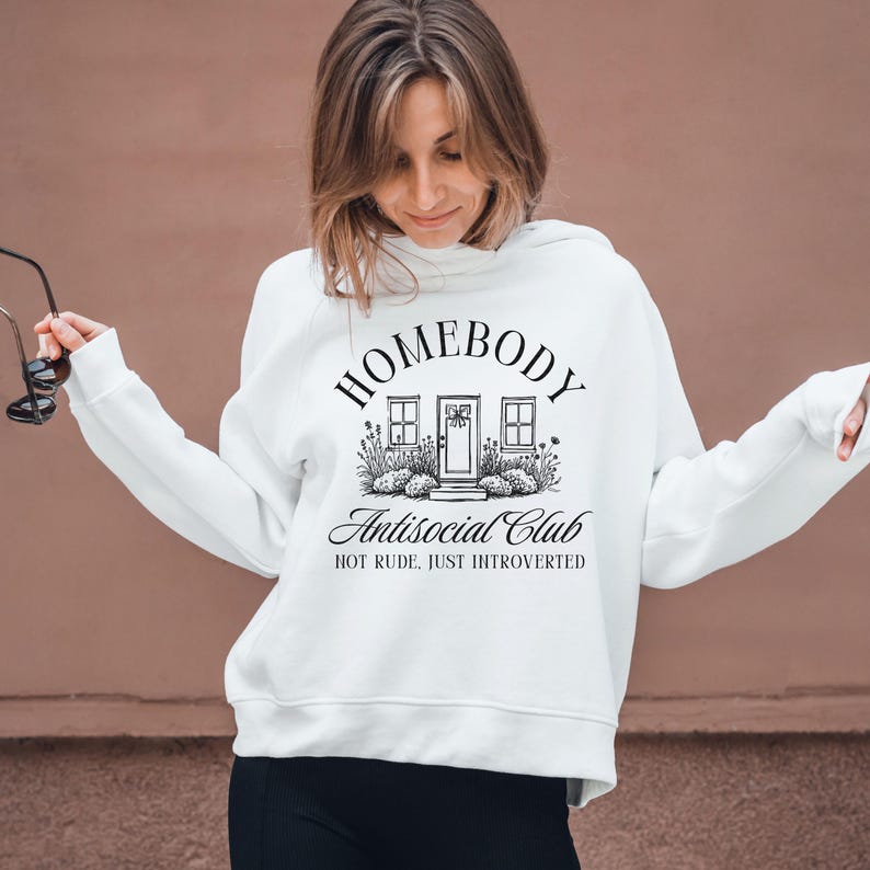 May include: White hoodie featuring the words "HOMEBODY Antisocial Club" in black script. The design includes a house graphic with windows and a door, surrounded by plants. The phrase "NOT RUDE, JUST INTROVERTED" is below the main text.
