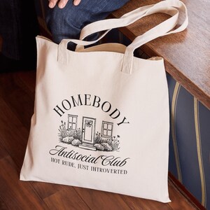 May include: A cream-coloured tote bag featuring black text and a house graphic. The text reads "HOMEBODY Antisocial Club HOT RUDE, JUST INTROVERTED". The bag has two handles and is made of a durable fabric.