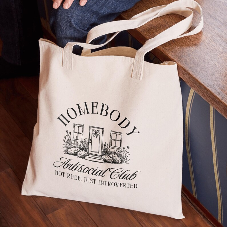 May include: A cream-coloured tote bag featuring black text and a house graphic. The text reads "HOMEBODY Antisocial Club HOT RUDE, JUST INTROVERTED". The bag has two handles and is made of a durable fabric.