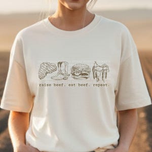 May include: Cream-colored t-shirt featuring a line drawing of a steak, cowboy boots, a hamburger, and a saddle. The text "raise beef. eat beef. repeat." is printed below the illustrations. The shirt is made of a soft material.