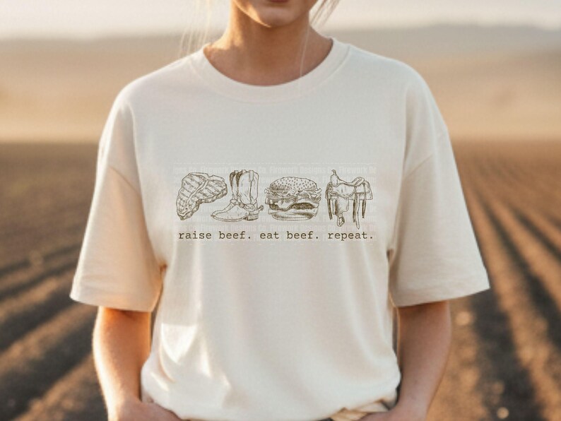 May include: Cream-colored t-shirt featuring a line drawing of a steak, cowboy boots, a hamburger, and a saddle. The text "raise beef. eat beef. repeat." is printed below the illustrations. The shirt is made of a soft material.