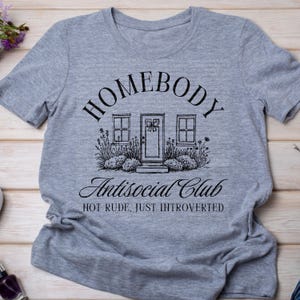 May include: A grey t-shirt with the text "HOMEBODY Antisocial Club" in black script. The graphic shows a house illustration with a door and windows. The words "NOT RUDE, JUST INTROVERTED" are below the graphic.