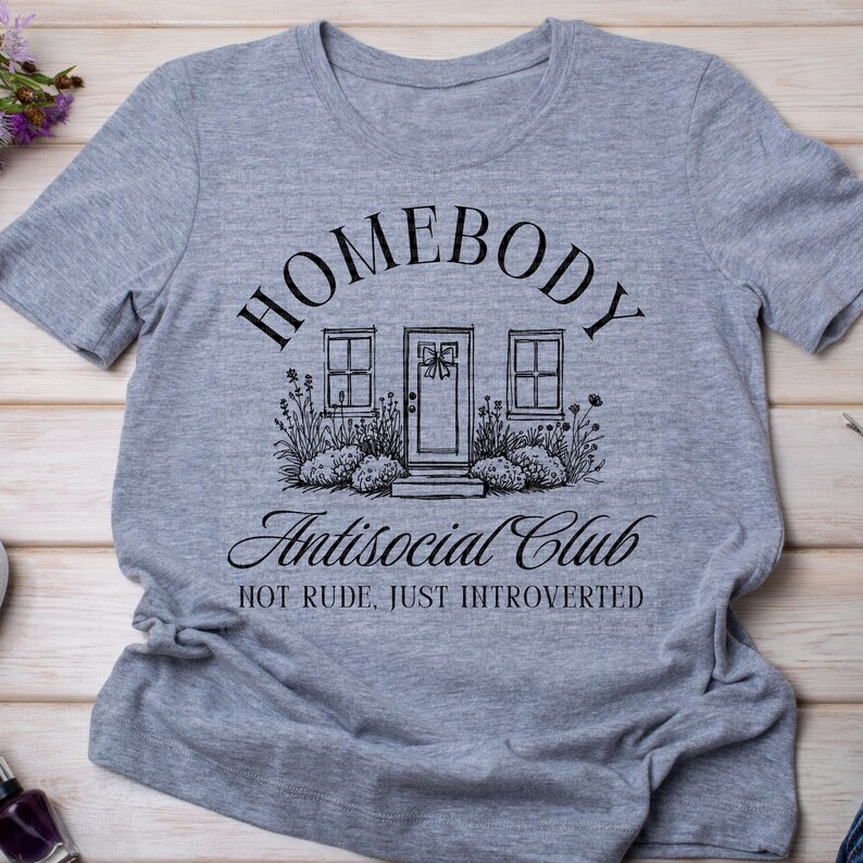May include: A grey t-shirt with the text "HOMEBODY Antisocial Club" in black script. The graphic shows a house illustration with a door and windows. The words "NOT RUDE, JUST INTROVERTED" are below the graphic.
