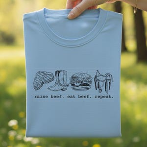 May include: A light blue t-shirt with a crew neck, featuring a black and white graphic of a steak, cowboy boots, a hamburger, and a saddle. The text "raise beef. eat beef. repeat." is printed below the graphics.