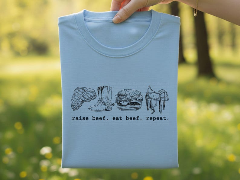 May include: A light blue t-shirt with a crew neck, featuring a black and white graphic of a steak, cowboy boots, a hamburger, and a saddle. The text "raise beef. eat beef. repeat." is printed below the graphics.