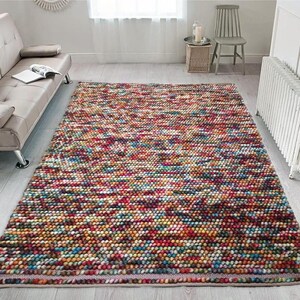 May include: A rectangular area rug with a textured, multi-colored design. The rug features a dense pattern of small, round, colorful elements in shades of red, orange, yellow, blue, and green. The rug is placed on a light-colored wooden floor.