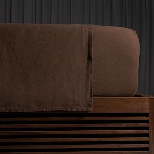 May include: A close-up of a bed with a brown linen bedspread and a wooden bed frame. The bedspread is a solid, dark brown color and appears to be made of a soft, textured fabric. The bed frame has a slatted design.