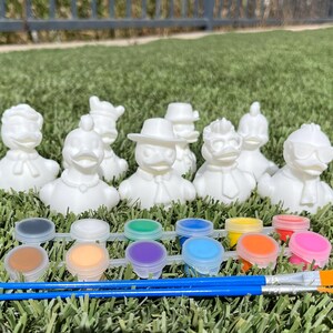 May include: A set of white ceramic duck figurines with various outfits, ready to be painted. Includes a paint palette with multiple colors and two blue paintbrushes. The items are arranged on a bed of green grass.