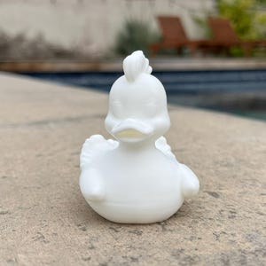 May include: A small, white, ceramic duck figurine with a detailed design. The duck has a crest on its head, wings, and a rounded body. It is sitting on a concrete surface, with a pool and greenery in the background.