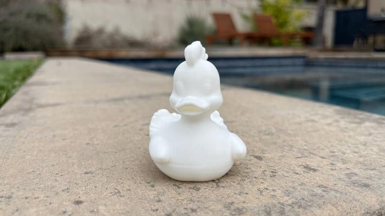 May include: A small, white, ceramic duck figurine with a detailed design. The duck has a crest on its head, wings, and a rounded body. It is sitting on a concrete surface, with a pool and greenery in the background.