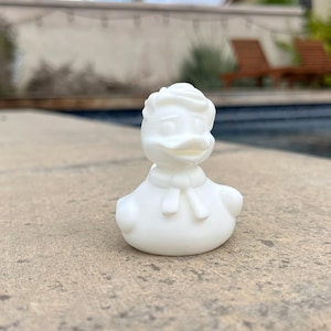 May include: A white rubber duck figurine, styled after a cartoon character, with a distinctive hairstyle and bow tie. The duck is positioned on a stone surface, with a swimming pool and greenery in the background.