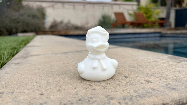 May include: A white rubber duck figurine, styled after a cartoon character, with a distinctive hairstyle and bow tie. The duck is positioned on a stone surface, with a swimming pool and greenery in the background.