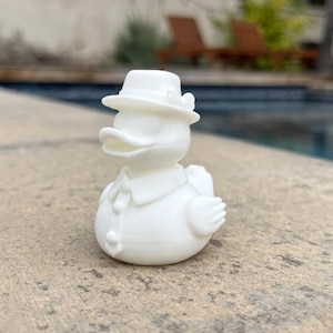 May include: A white duck figurine wearing a hat, shirt, and tie. The duck is standing on a concrete surface with a pool in the background. The duck has a classic, vintage design.