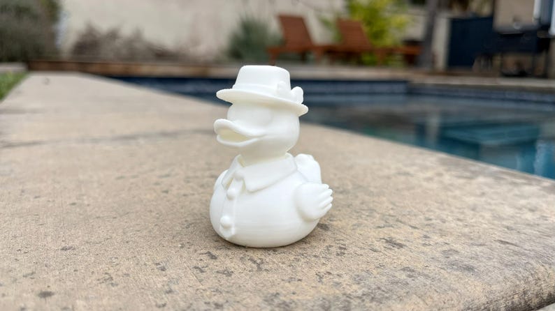 May include: A white duck figurine wearing a hat, shirt, and tie. The duck is standing on a concrete surface with a pool in the background. The duck has a classic, vintage design.