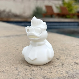 May include: A white rubber duck figurine with a stylish design. The duck is wearing glasses and a scarf. The duck is sitting on a concrete surface near a pool.