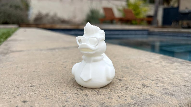 May include: A white rubber duck figurine with a stylish design. The duck is wearing glasses and a scarf. The duck is sitting on a concrete surface near a pool.