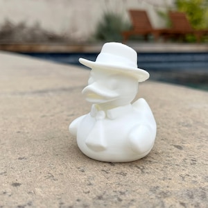 May include: A white rubber ducky wearing a white fedora hat, a bow tie, and a collared shirt. The duck is sitting on a concrete surface, with a swimming pool and greenery in the background.