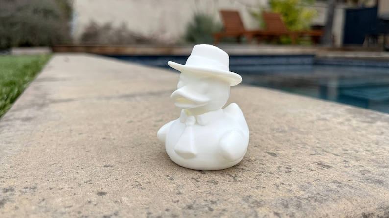 May include: A white rubber ducky wearing a white fedora hat, a bow tie, and a collared shirt. The duck is sitting on a concrete surface, with a swimming pool and greenery in the background.