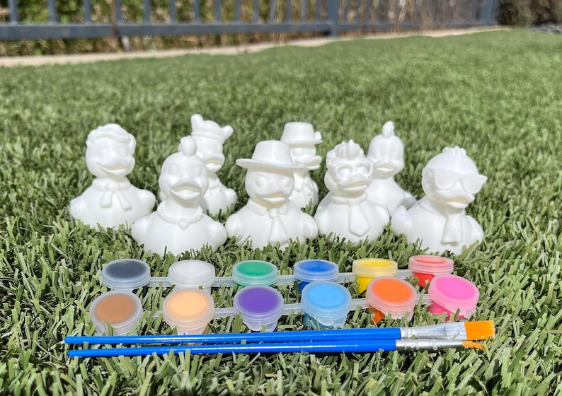 May include: A set of white ceramic duck figurines with various outfits, ready to be painted. Includes a paint palette with multiple colors and two blue paintbrushes. The items are arranged on a bed of green grass.