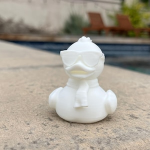 May include: A white rubber duck wearing sunglasses and a scarf. The duck is positioned on a stone surface with a swimming pool in the background. The duck has a classic rubber duck shape.