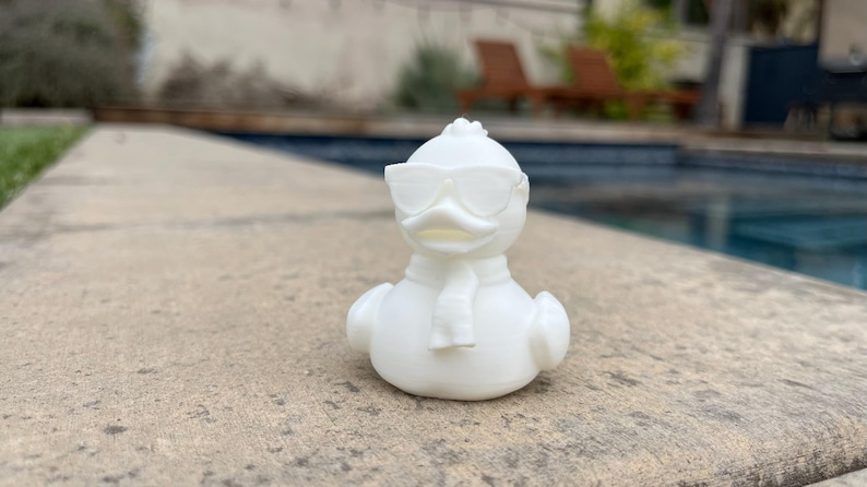 May include: A white rubber duck wearing sunglasses and a scarf. The duck is positioned on a stone surface with a swimming pool in the background. The duck has a classic rubber duck shape.