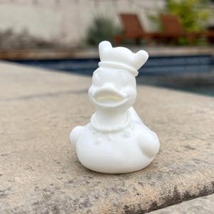 May include: A white ceramic duck figurine with a chef's hat and a beaded necklace. The duck is sitting on a stone surface, with a swimming pool in the background.