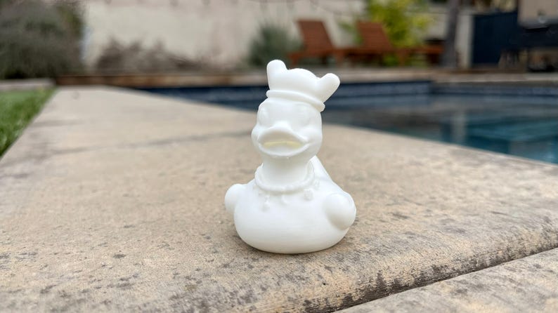 May include: A white ceramic duck figurine with a chef's hat and a beaded necklace. The duck is sitting on a stone surface, with a swimming pool in the background.