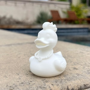 May include: A white decorative duck figurine with a pearl necklace and a floral headpiece. The duck is sitting on a concrete surface near a swimming pool. The duck has a detailed design.