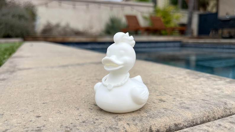 May include: A white decorative duck figurine with a pearl necklace and a floral headpiece. The duck is sitting on a concrete surface near a swimming pool. The duck has a detailed design.