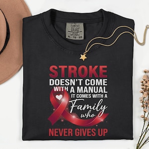 May include: Black t-shirt with the text "STROKE DOESN'T COME WITH A MANUAL IT COMES WITH A Family who NEVER GIVES UP" in red and white. A red ribbon graphic is featured. A gold necklace with a star pendant is draped on the shirt.