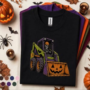 May include: A black crew neck t-shirt featuring a graphic of a skeleton wearing an orange hard hat operating a green and purple skid steer loader. The loader bucket is orange and decorated with a black jack-o'-lantern face design.