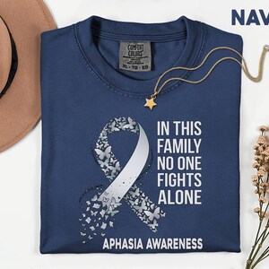 Może przedstawiać: Granatowa koszulka z białym wzorem wstążki z motyli oraz napisem "IN THIS FAMILY NO ONE FIGHTS ALONE" i "APHASIA AWARENESS". Złoty naszyjnik z zawieszką w kształcie gwiazdy leży na koszulce.