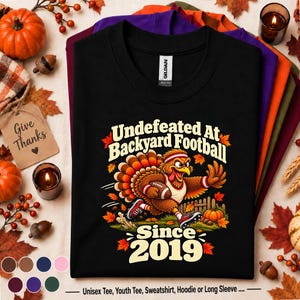 May include: A black t-shirt featuring a graphic of a turkey wearing a headband and jersey while running with a football. The text reads Undefeated At Backyard Football Since 2019. The design includes autumn leaves and a wooden fence background.
