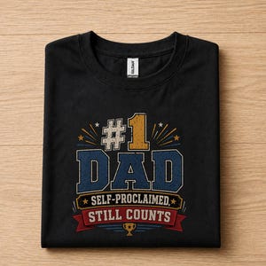 May include: Black t-shirt with the text "#1 DAD SELF-PROCLAIMED, STILL COUNTS" in a distressed, vintage style. The design includes gold, blue, and red accents, with a trophy graphic at the bottom. The shirt is folded neatly on a wooden surface.