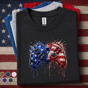 May include: Black t-shirt featuring a graphic of a video game controller in red, white, and blue, resembling the American flag. The controller design has a dripping paint effect. Several folded t-shirts in different colors are stacked behind the featured shirt.