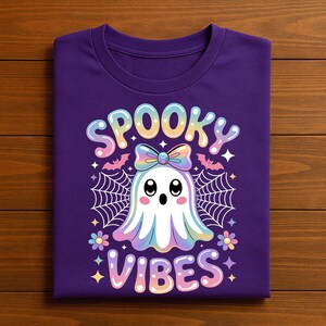 May include: A purple t-shirt featuring a cute ghost design with the words "Spooky Vibes". The ghost is white with a rainbow gradient, a bow, and is surrounded by spiderwebs, bats, and stars. The shirt is folded.