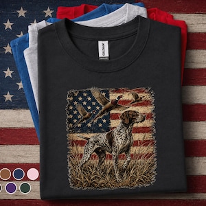 May include: A black crew neck t-shirt featuring a graphic print of a German Shorthaired Pointer standing in tall grass with two flying pheasants against a distressed American flag background. The shirt has a Gildan brand label at the inner neckline.
