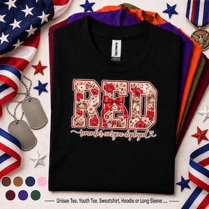 Puede incluir: Una camiseta negra con la palabra RED en letras grandes de bloque rellenas con un patrón floral de rosas rojas y pequeñas flores blancas. Un lazo rojo está atado alrededor de la letra central. Debajo, el texto dice remember everyone deployed.