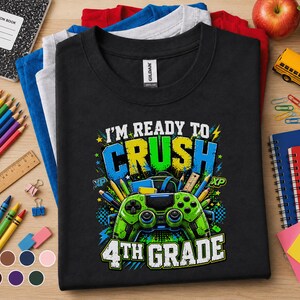 May include: A black short-sleeved t-shirt featuring a graphic print with the text "I'M READY TO CRUSH 4TH GRADE" in white and blue. The design includes a green video game controller, pencils, and school supplies in a vibrant, colorful style.