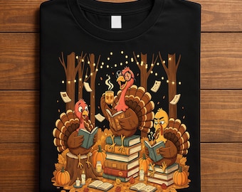 May include: Black t-shirt featuring a whimsical illustration of three turkeys reading books in a forest setting. The turkeys are surrounded by pumpkins, candles, and falling leaves, with string lights in the background. A cozy autumn-themed design.